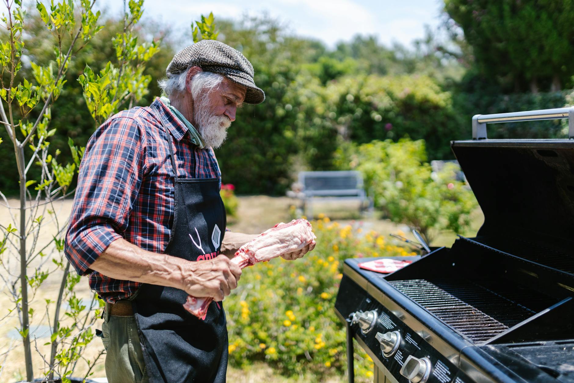 elderly man in plaid button up shirt standing in front of a griller holding meat